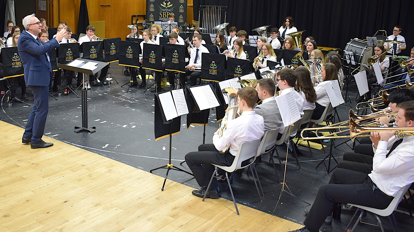 The National Youth Brass Band of Scotland during a performance conducted by Ian Porthouse. Young musicians sit in orchestral formation as Ian directs their performance.