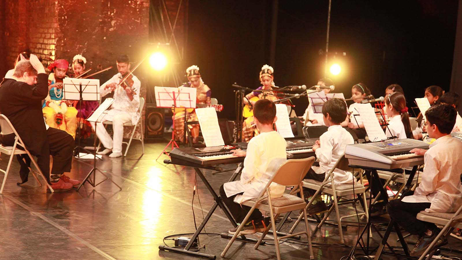 On a stage, a band of young musicians play together on keyboards and violins, wearing traditional Indian dress