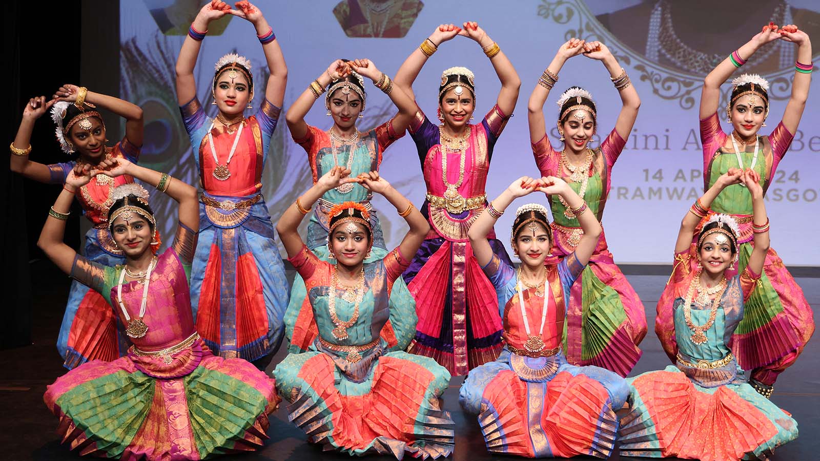 A group of young girls in traditional Indian dress pose together on a stage, smiling and with their hands clasped together in the air