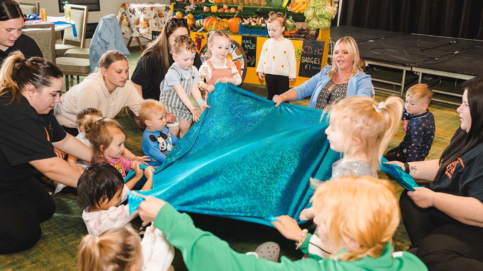 A group of nursery-aged children and adults all holding a shiny and sparkly blue blanket as they sing together