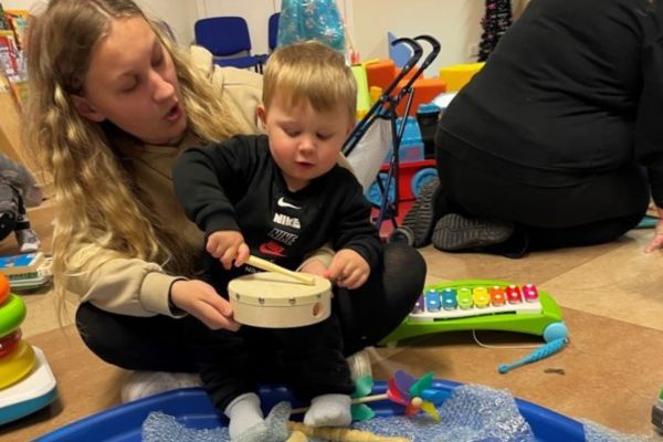 A mum sits with her child on her knee as they play with a small drum