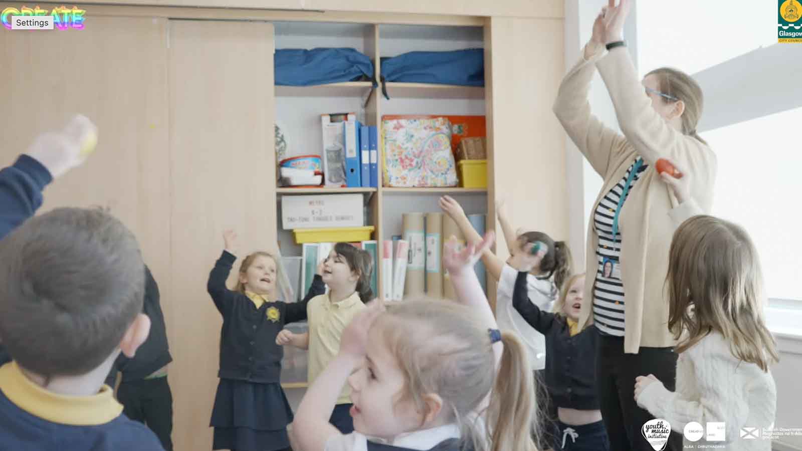 A classroom with primary-aged children and a teacher, signing and dancing together with their hands in the air.