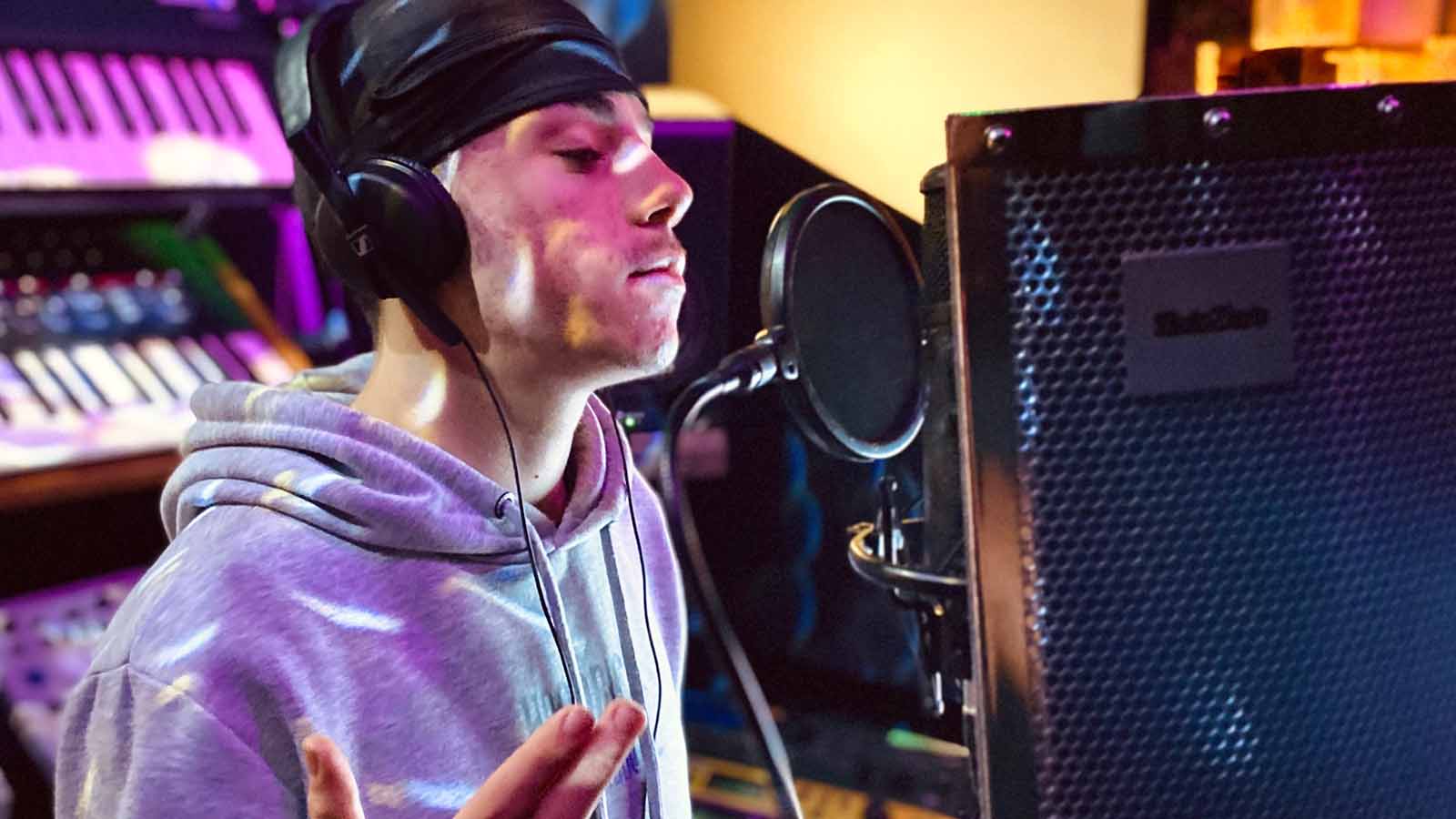 A man with headphones in a grey hoodie speaking or singing into a mic in a recording studio.