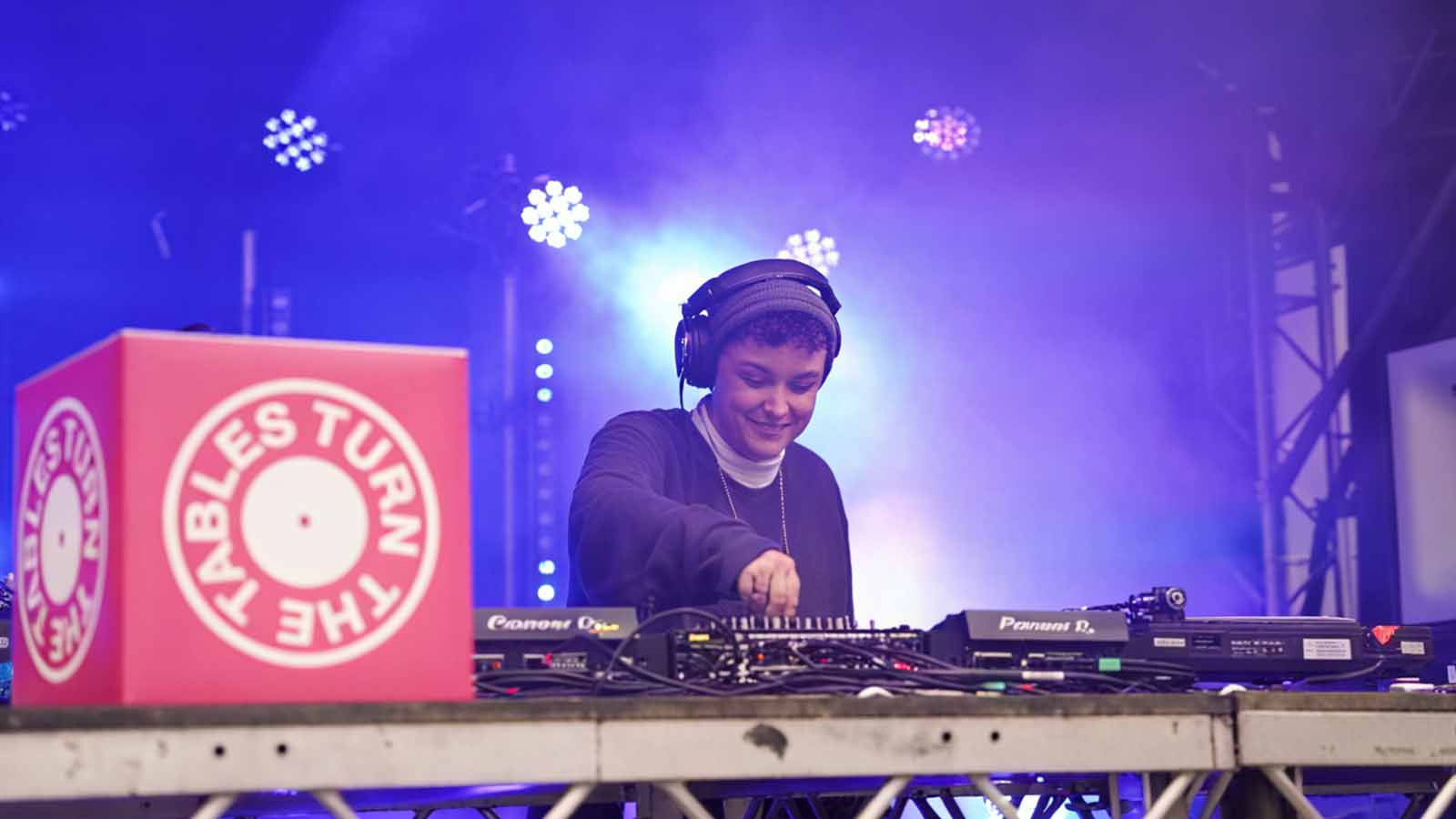 A young person working a DJ deck on a stage during an event, lit from behind by stage lights and with the Turn The Tables logo depicted in pink and white on a sign to the left.