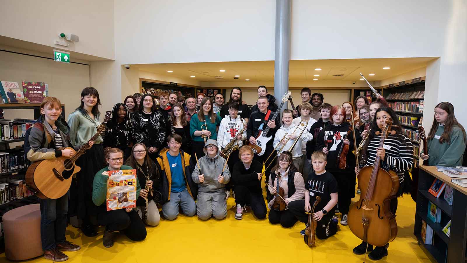 A large group of young people holding various musical instruments including string and wind instruments, posing with pop star and musician Ed Sheeran in a library with bright yellow floors and tall white walls