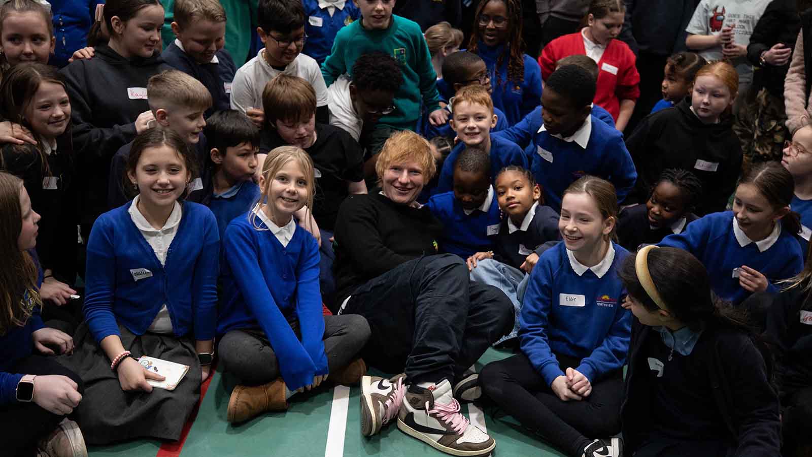 A group of primary school pupils in bright blue jumpers sitting on a gym floor smiling and posing with a man with red hair - this is Ed Sheeran.