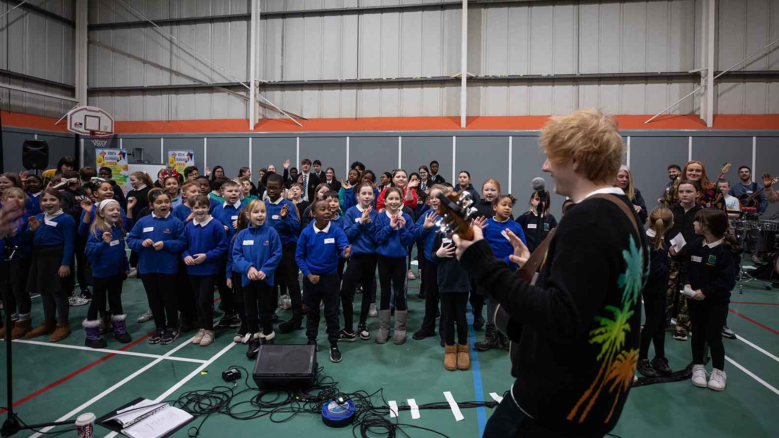 A group of primary school pupils in bright blue jumpers in a large gym hall, smiling and clapping along with a man with red hair - this is Ed Sheeran.