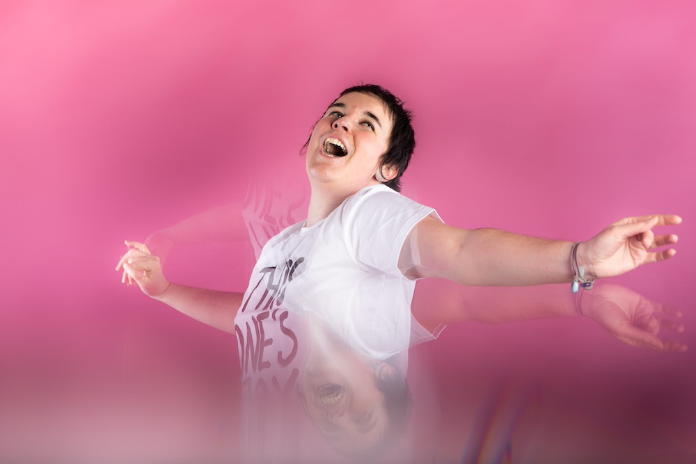 A woman dances joyfully in front of a pink background, smiling with her mouth open looking triumphant