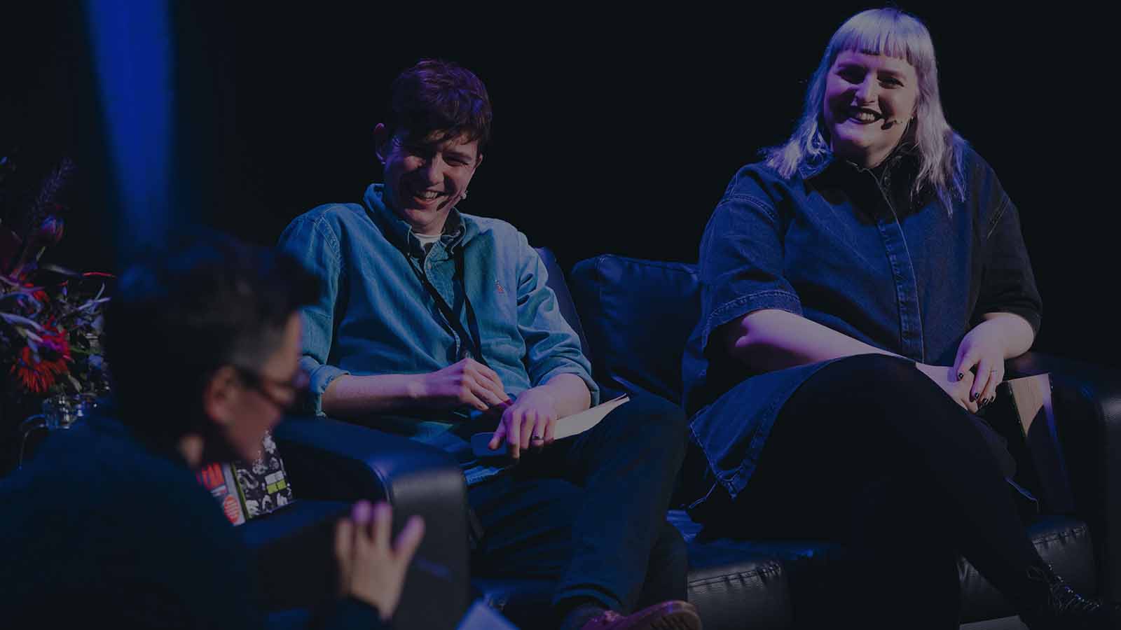 A man in a blue denim shirt with short brown hair and a woman with blonde hair and wearing dark lipstick - both wearing microphones, sit on a sofa listening to another person talking.