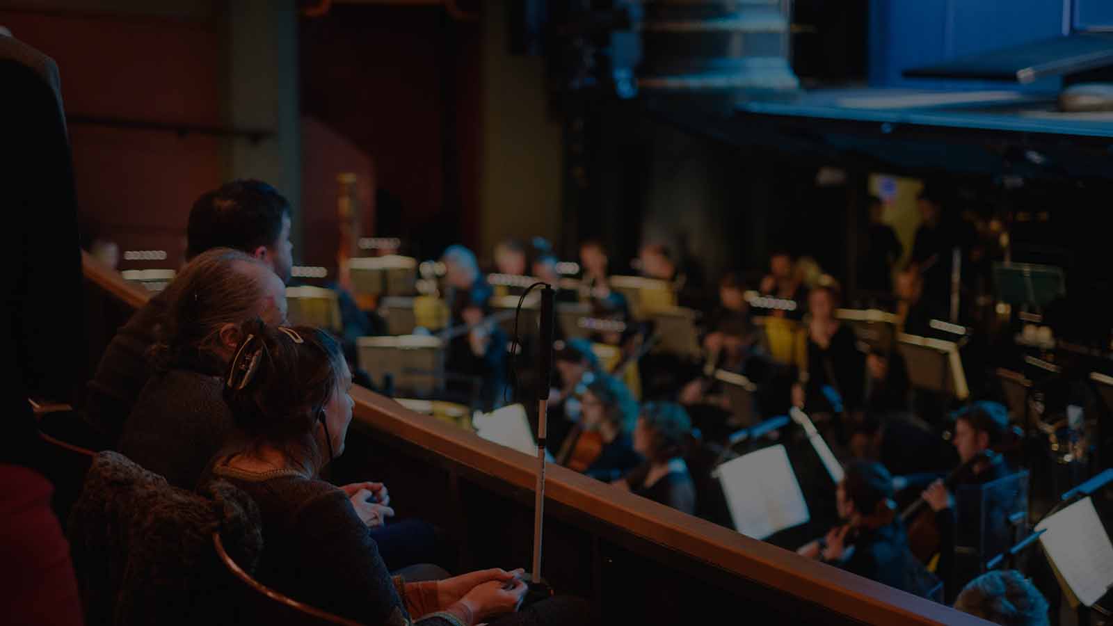 A recording studio - in the background and out of focus an orchestra sits playing music. In the foreground three people sit watching and listening - a woman with a hair clip and a guide cane, an older man with grey hair and another man with black hair.