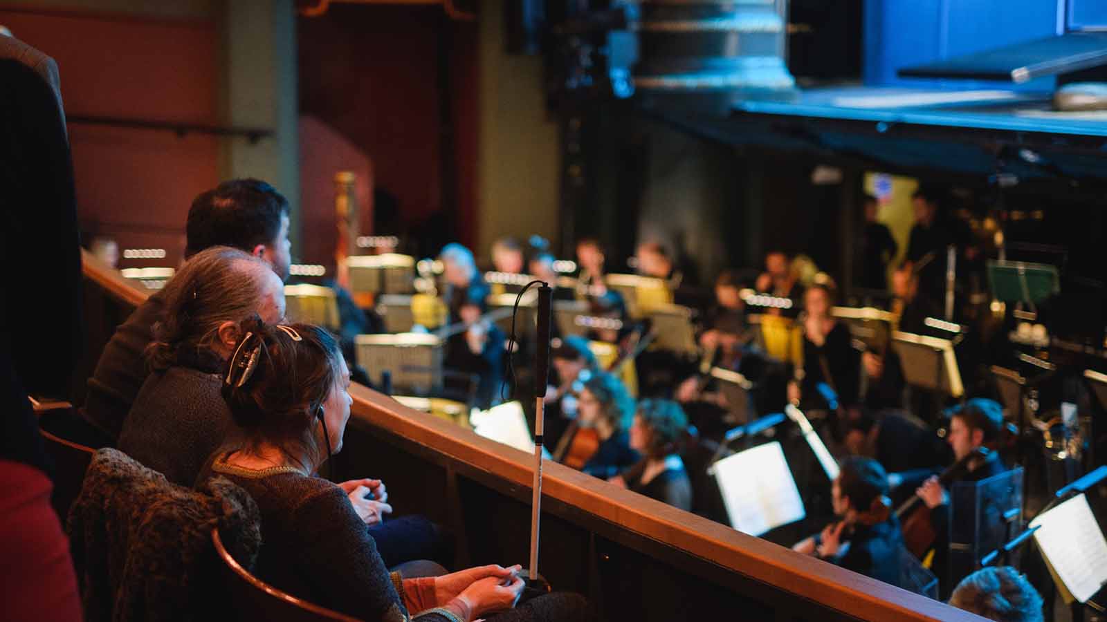 A recording studio - in the background and out of focus an orchestra sits playing music. In the foreground three people sit watching and listening - a woman with a hair clip and a guide cane, an older man with grey hair and another man with black hair.