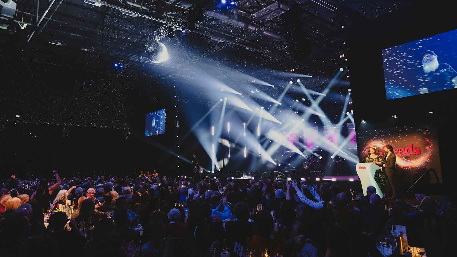 A large auditorium with a stage lit by many spotlights, refracting and reflecting off a disco ball suspended from the ceiling. A large audience crowded round tables watch as two people stand at a lectern - this is the Na Trads ceremony.