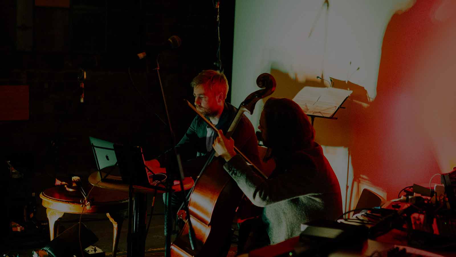 In a small space lit by moody orange and white lights, a man sits at a laptop working while next to him a woman holding a Cello watches, they appear to be working on music together. They’re surrounded by sheet music on stands, decks, tables and mics.