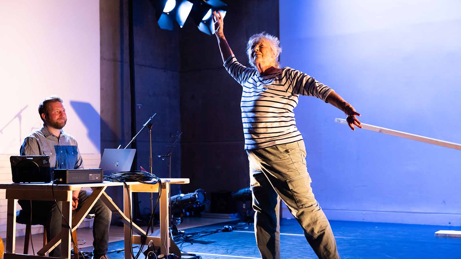 A stage with two large white projector screens. In front of one is a man sitting at a table with a laptop and what appears to be lighting or sound decks. A woman in a striped jumper and cargo trousers holds her arms up in a pose, as if she’s dancing.