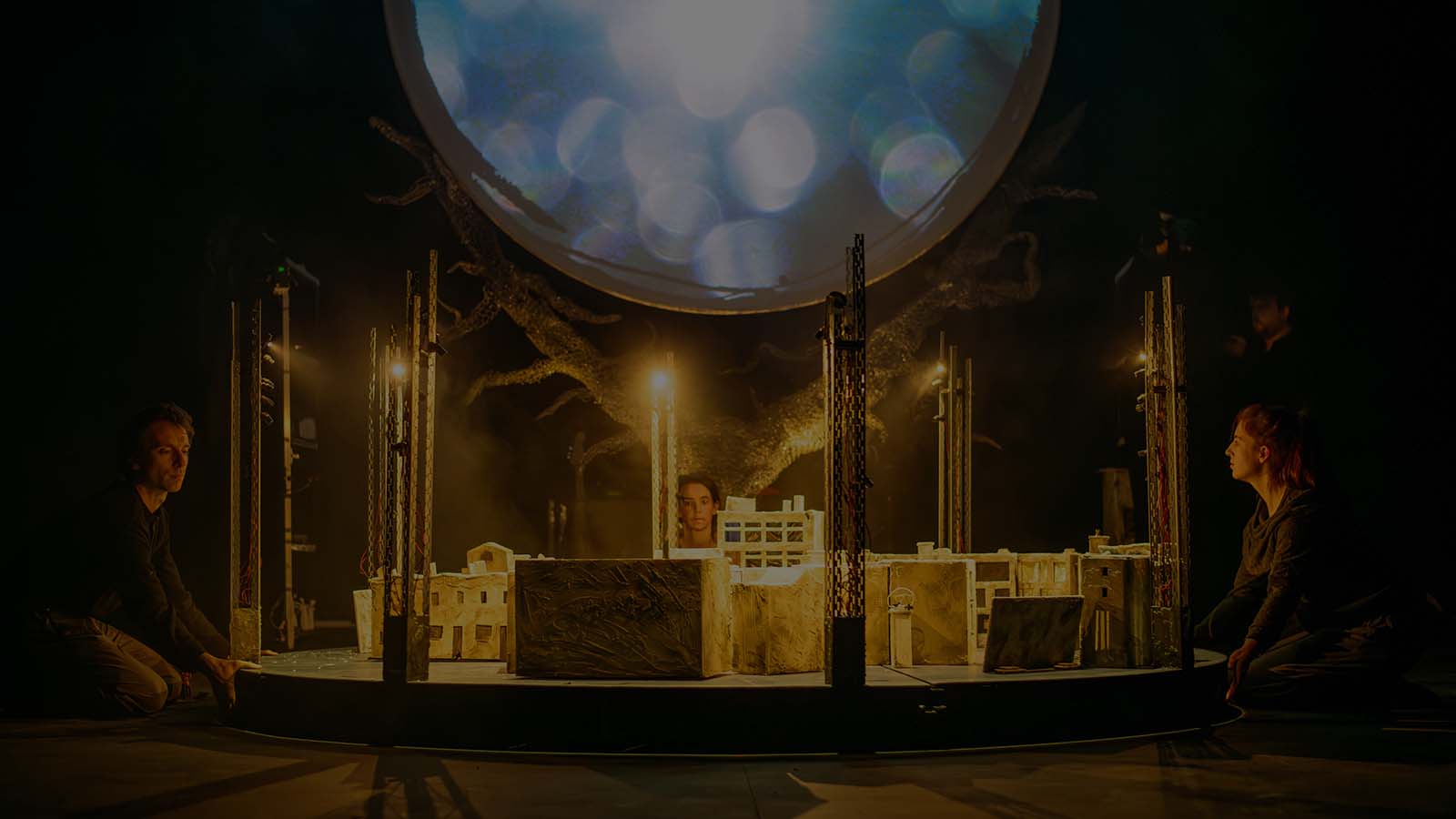 A stage; on it three people kneel around a slightly raised dais on which sits a while, detailed, paper mache city, softly and warmly lit by floodlights. In the middle, a large circle of sparking white light, seemingly held up by a leafless tree.