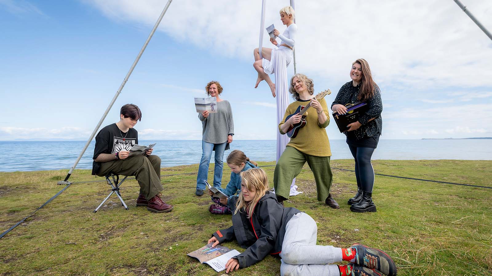 A group of people pictured in front of a coastal view are reading the Findhorn Bay Festival's 2024 programme. One is a performer suspended from white cloth ropes, one is playing the ukulele, others are sitting, standing and lying down.