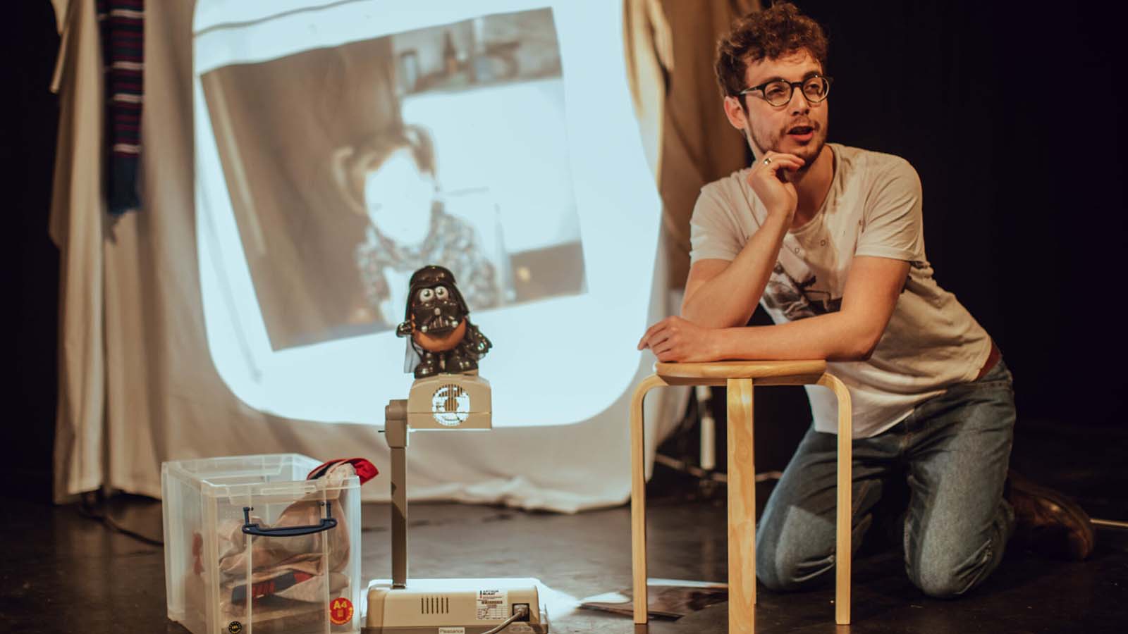 A man in a white t-shirt and jeans leans on a small wooden stool, his chin resting in his right hand. Next to him on the right is an old projector, which casts a blurry childhood photo on the back of the stage. This is a still from Jack Hunter's play One of Two.