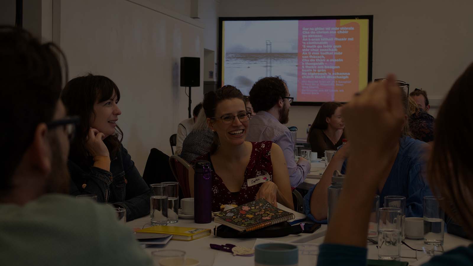 A group of people sitting and chatting together at a table, covered in notebooks, water glasses and cups of tea. They are in a large room, where more tables can be seen in the background, and a colourful image on a screen behind them in written in Scottish Gaelic.