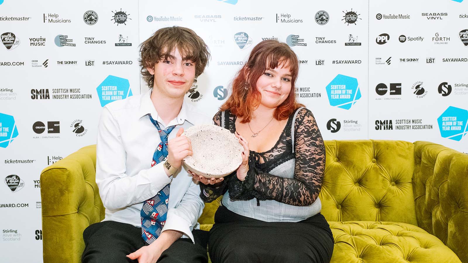 A young man with long brown hair in a white shirt and colourful graphic tie, and a young woman with short pink hair in a black lace dress and patterned top holding a sculpture as they sit together on a bright yellow velvet sofa. These are the members of No Windows with their Sound of Young Scotland Award 2023.