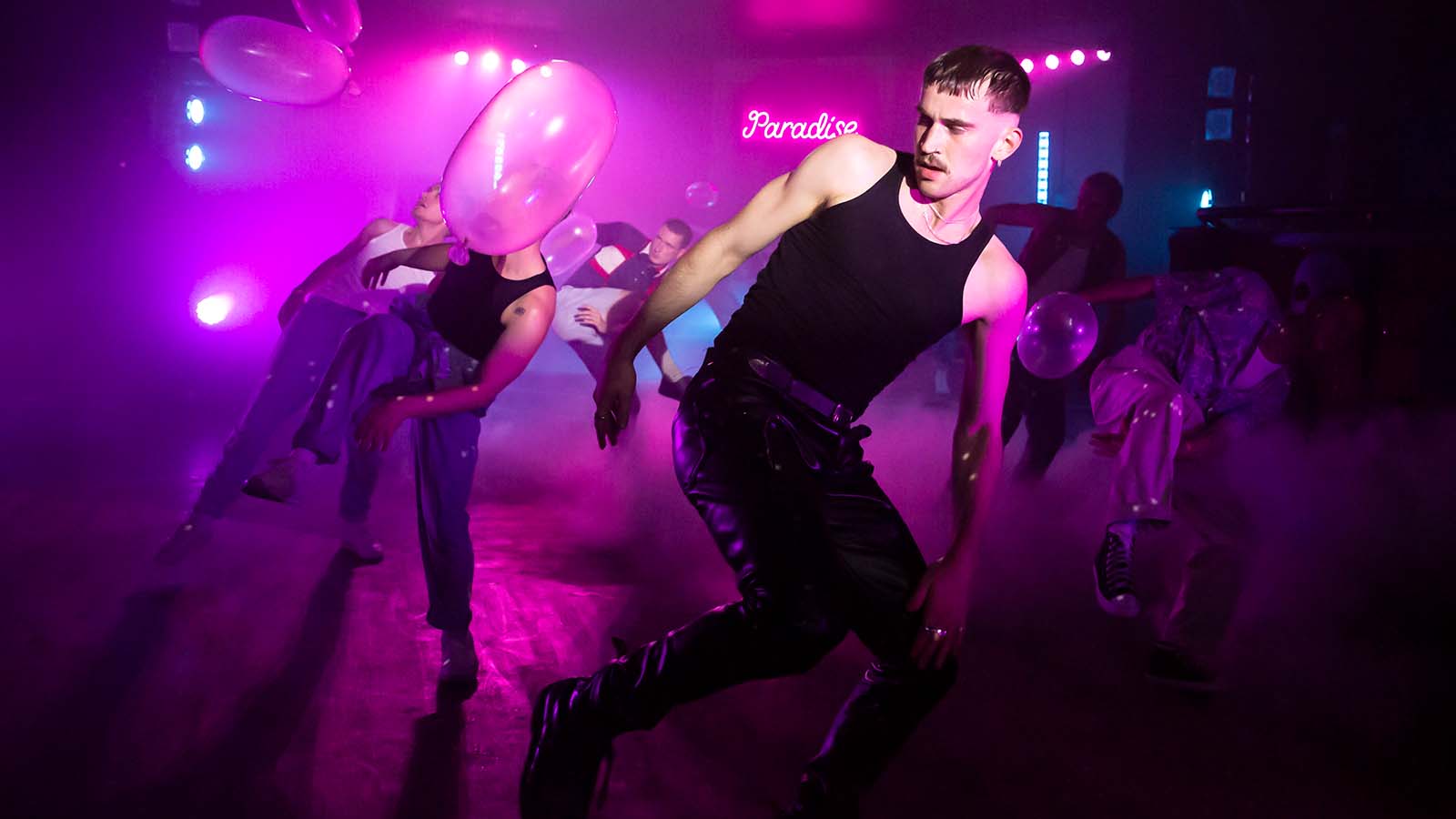 A group of dancers partying on a stage lit in smokey pink and purple lights. Balloons fall from the ceiling and in the background, the word 'Paradise' is lit up in pink neon light.