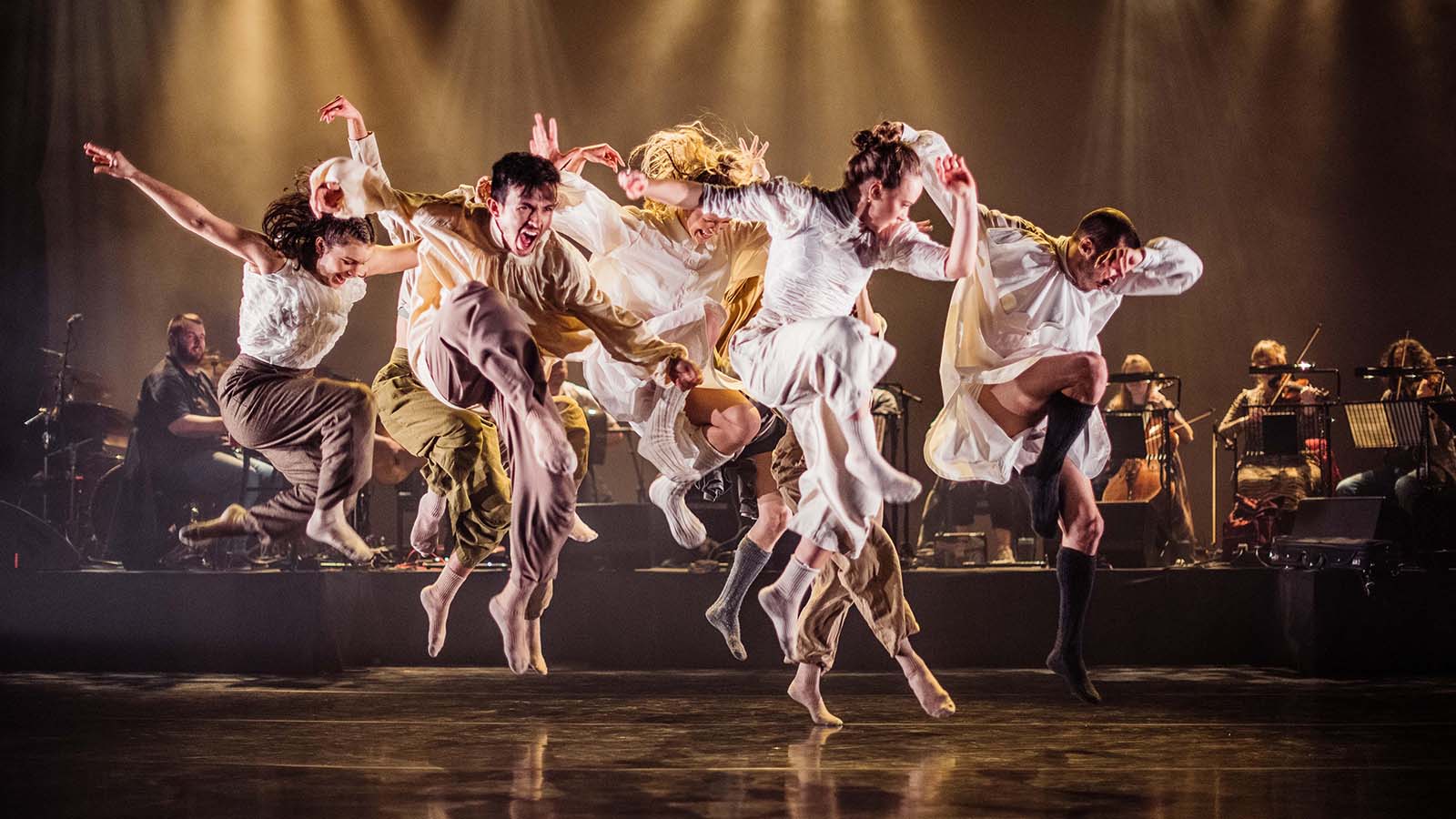 A group of dancers leaping in the air during a performance. Behind them is a live orchestra.