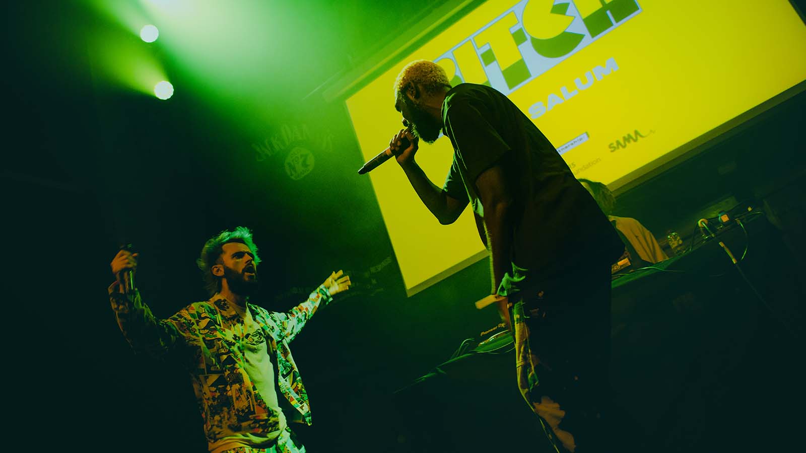 Two performers on stage during a Pitch Scotland gig - their logo is displayed at the back of the stage and moody green lighting fills the space