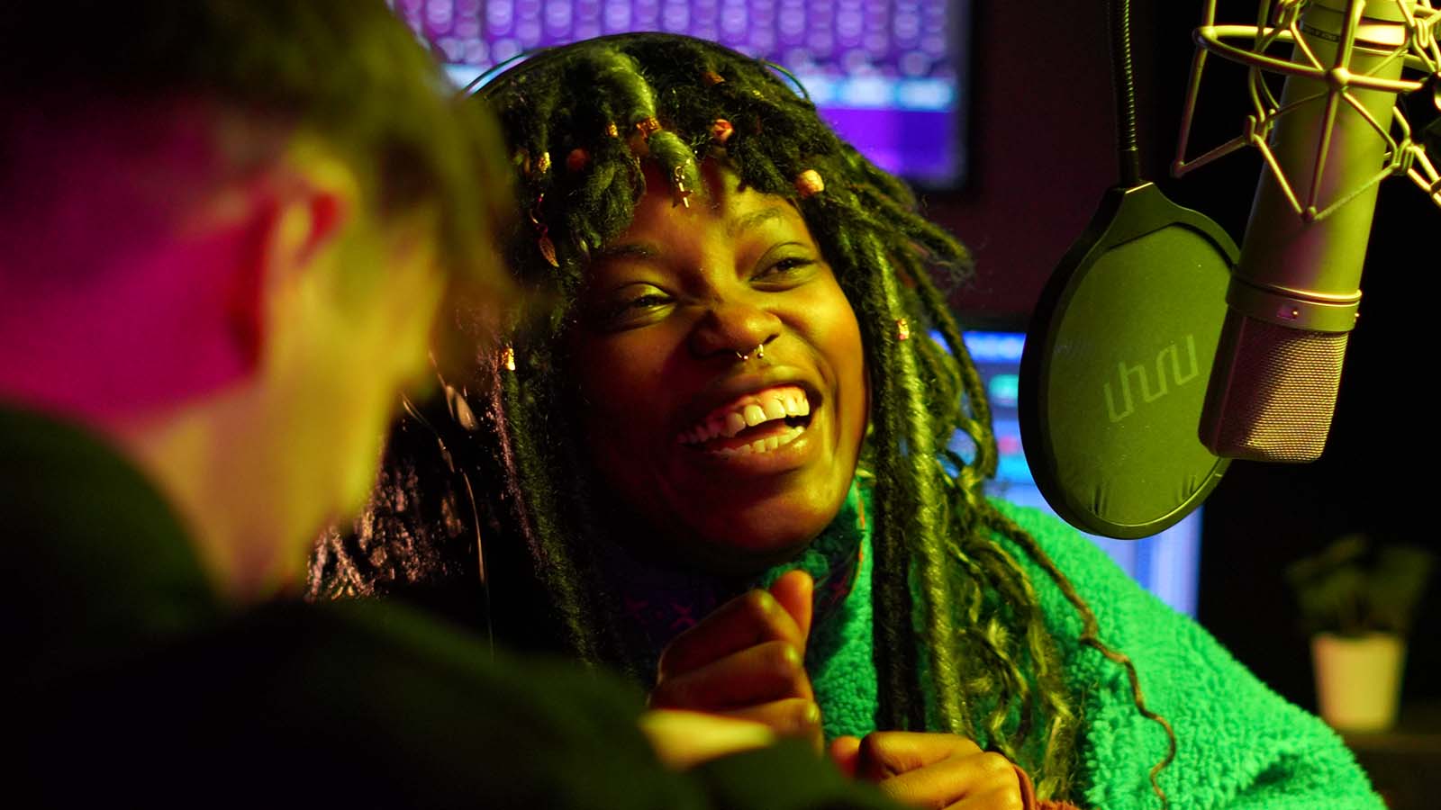 A young Black woman smiling, she is in a recording studio, with a professional microphone just in front of her