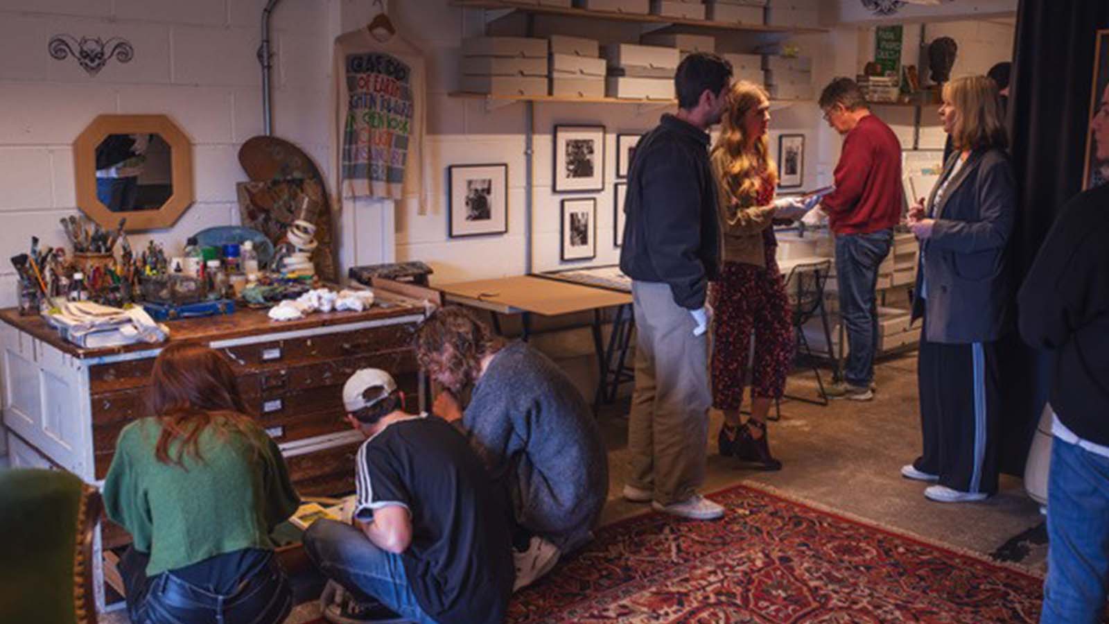 Several people exploring a small storage room with art supplies, desks and equipment, a patterned Persian rug, boxes on high shelves and framed professional black and white photos on the wall.
