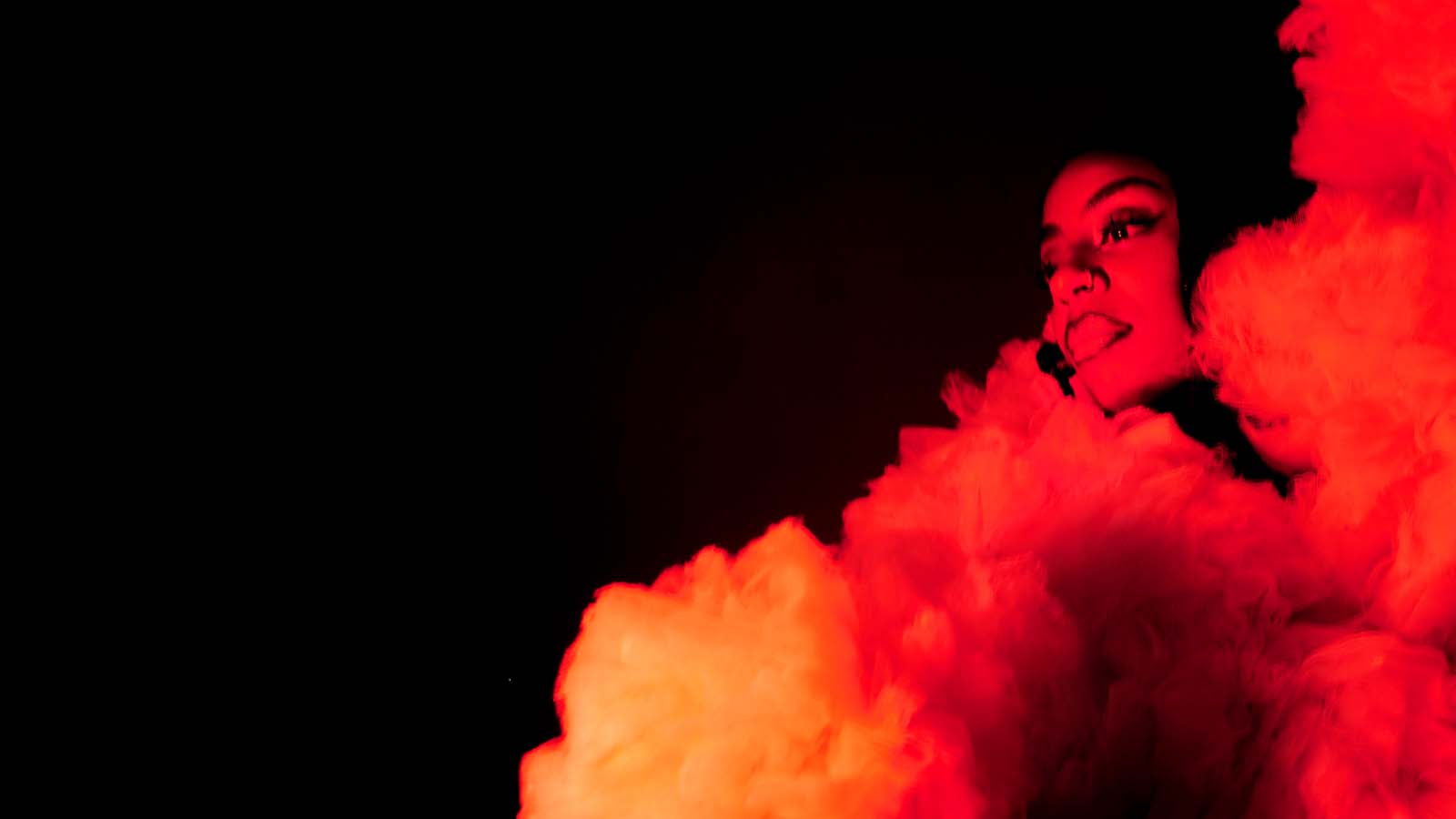 A woman (Male Broomes) surrounded by clouds of what appears to be smoke or cloth, lit in dramatic red and white lighting