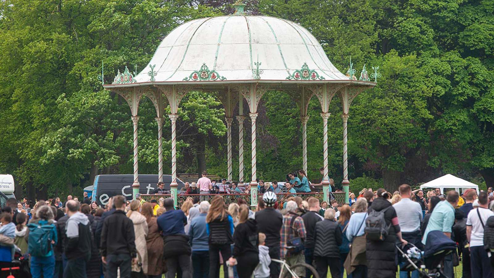 A large crowd in a green and leafy park stand watching a band perform on a gazebo