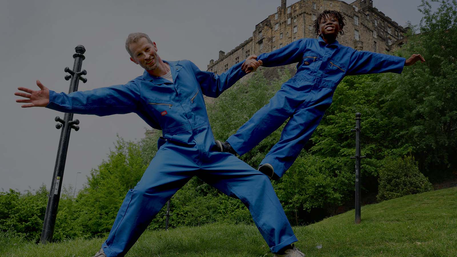 A man and a boy in bright blue jumpsuits in a garden. They are dancing joyfully with each other, striking a pose with the boy standing on the mans legs as they stay connected by holding hands.