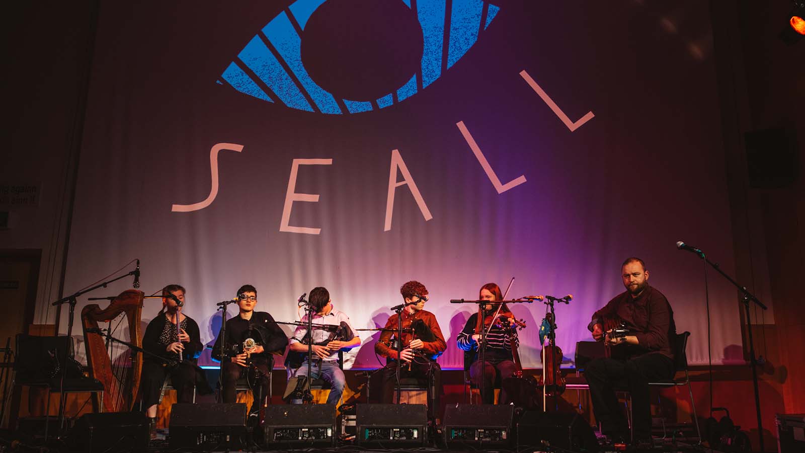 A group of young musicians seated in a row on a stage playing various instruments: a flute, pipes, fiddles - at the end of the row is a man playing the guitar. On the wall behind them a giant projection reads 'SEALL'