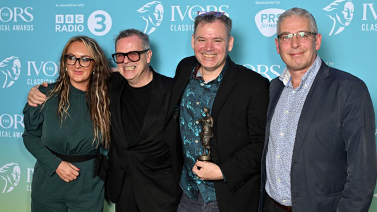 Four people, a woman and three men standing together smiling and hugging as they hold a small bronze statue - this is the Red Note Ensemble/Dumbworld team who have just accepted the Ivor Novello Classical Award for their Easterhouse Children’s Manifesto project.