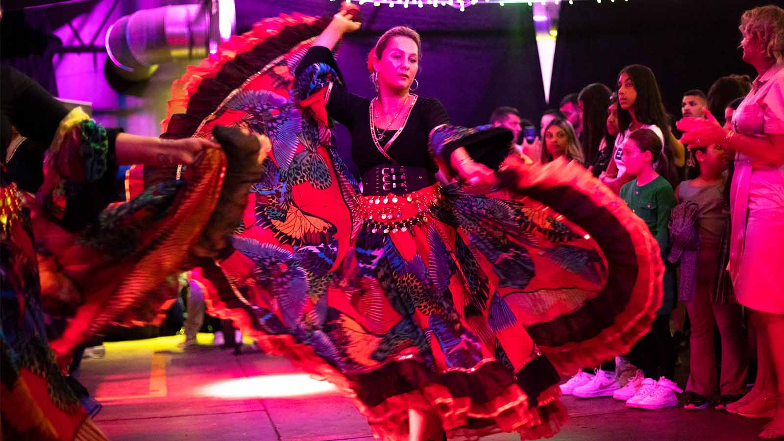 A woman dances in a bright and colourful black and patterned dress decorated with small silver buckles and other details. She is holding the ends of her large long and flowing skirt as she twirls before a crowd of families and children.