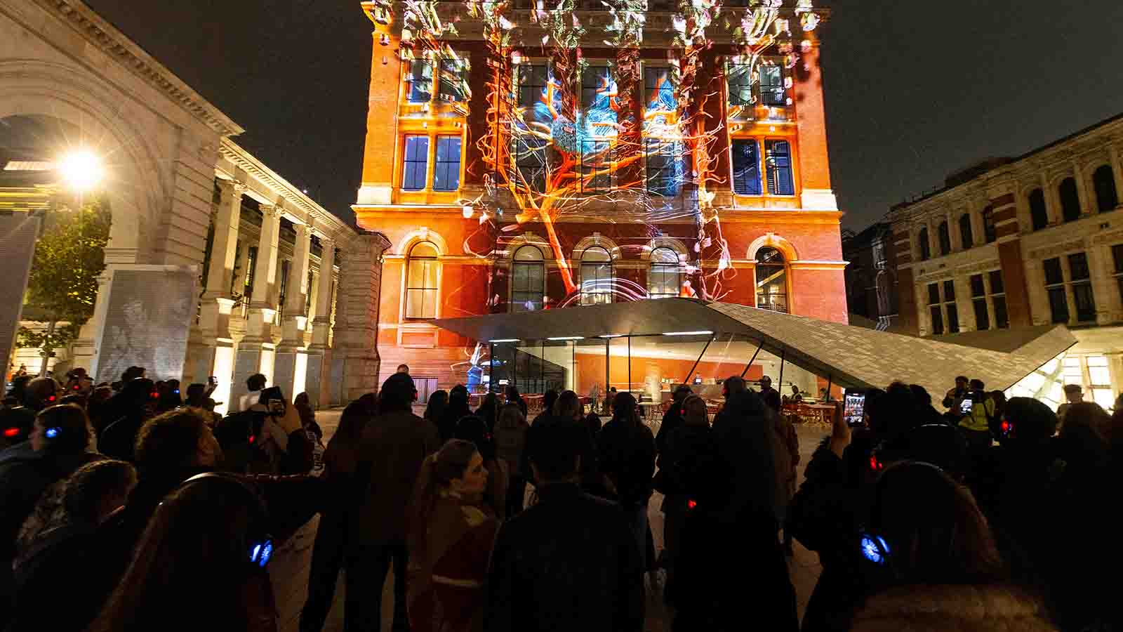 A crowd of people gathered on a dark evening in winter watching a light projection on an old building.