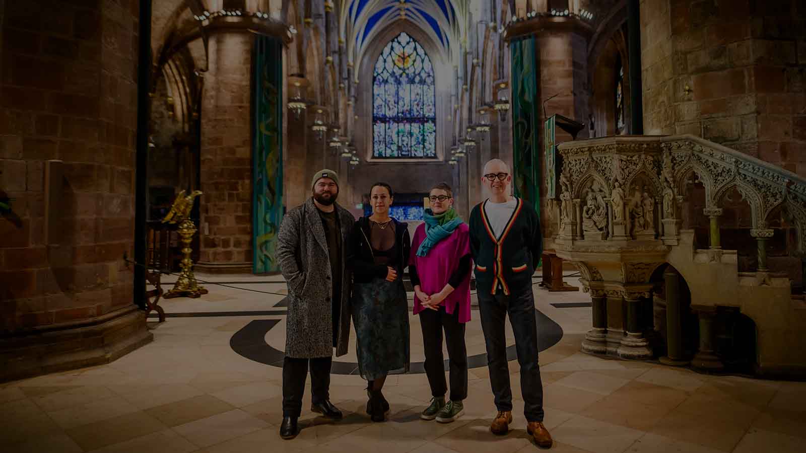 Four people (a man in a green beanie and long grey coat, a woman in a blue skirt and black jacket and very short black hair, a woman in a bright pink jumper and blue/green scarf with short dark hair, a man with shirt white hair and black-rimmed glasses wearing a white t-shirt, jeans and a cardigan) stand together inside a stone large cathedral building, a highly detailed pulpit to their right and golden lectern to the their left, and an intricate stained glass window pictured at the very back.