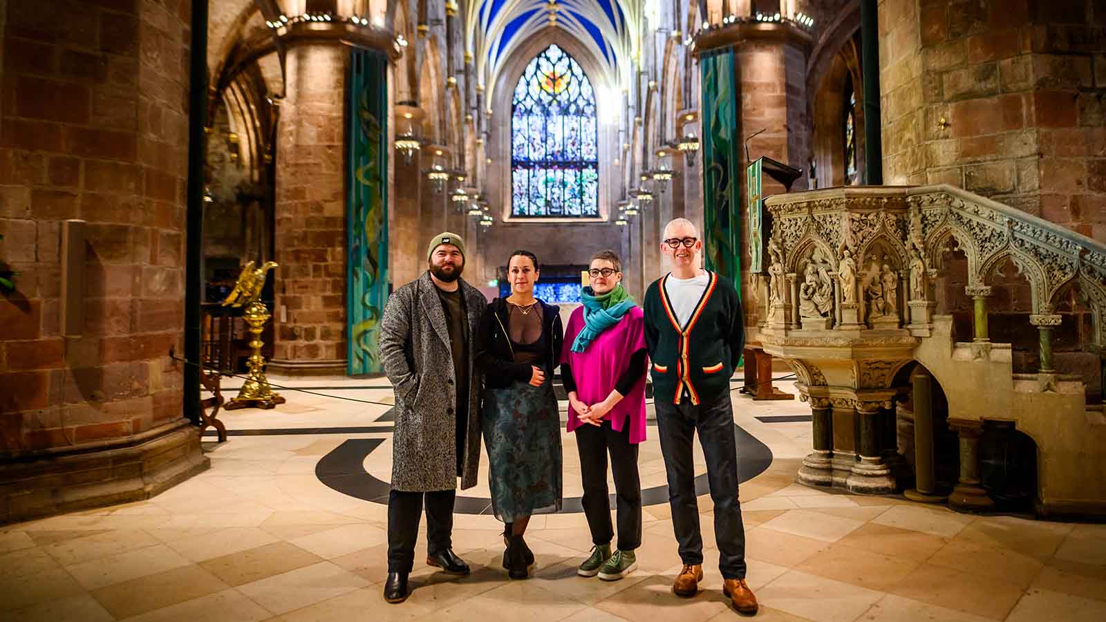 Four people (a man in a green beanie and long grey coat, a woman in a blue skirt and black jacket and very short black hair, a woman in a bright pink jumper and blue/green scarf with short dark hair, a man with shirt white hair and black-rimmed glasses wearing a white t-shirt, jeans and a cardigan) stand together inside a stone large cathedral building, a highly detailed pulpit to their right and golden lectern to the their left, and an intricate stained glass window pictured at the very back.