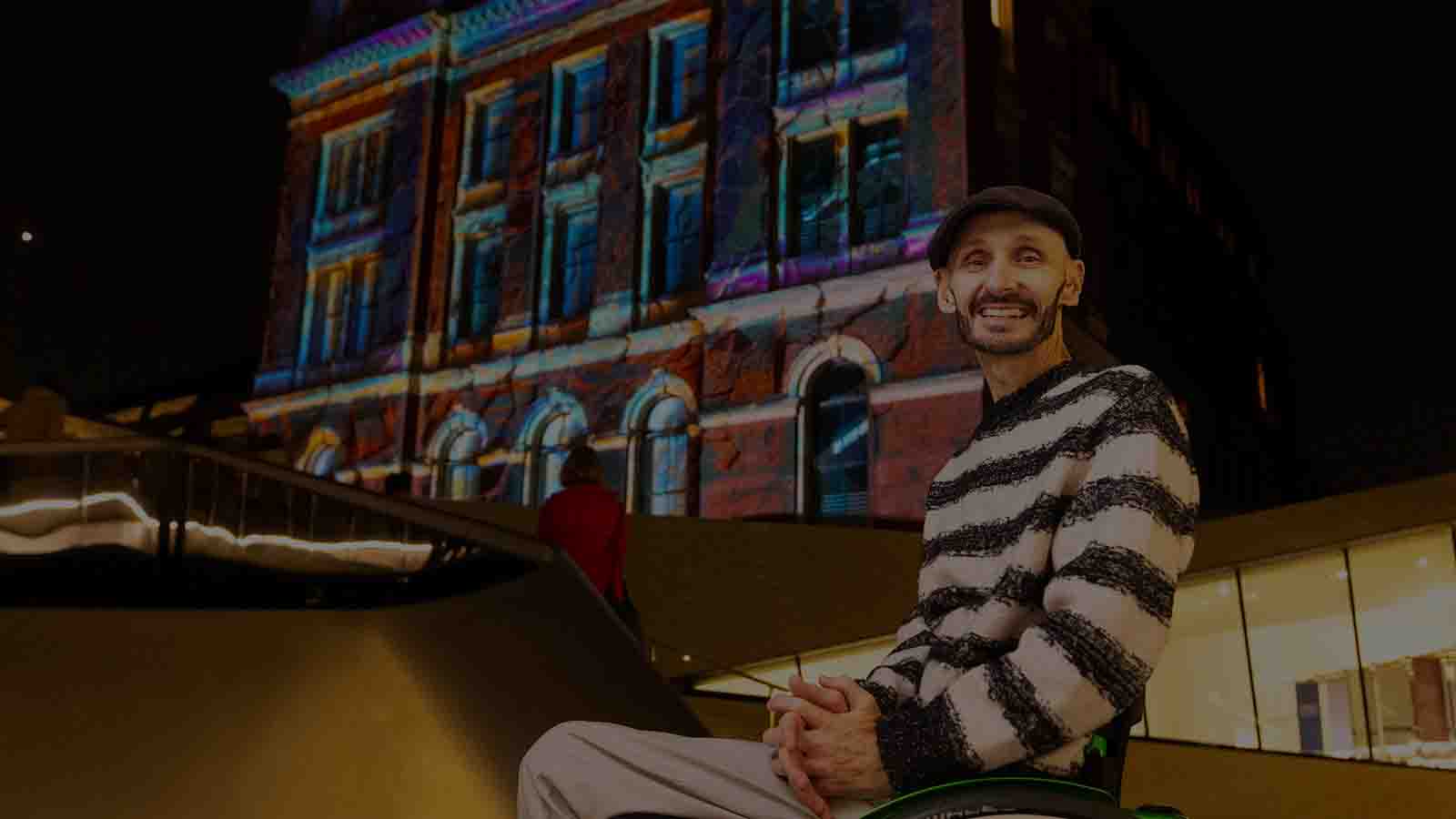 A man in a black flat cap and black and white striped jumper in a wheelchair (choreographer and producer Marc Brew) pictured in front of an old building with a colourful light show projected on to it.