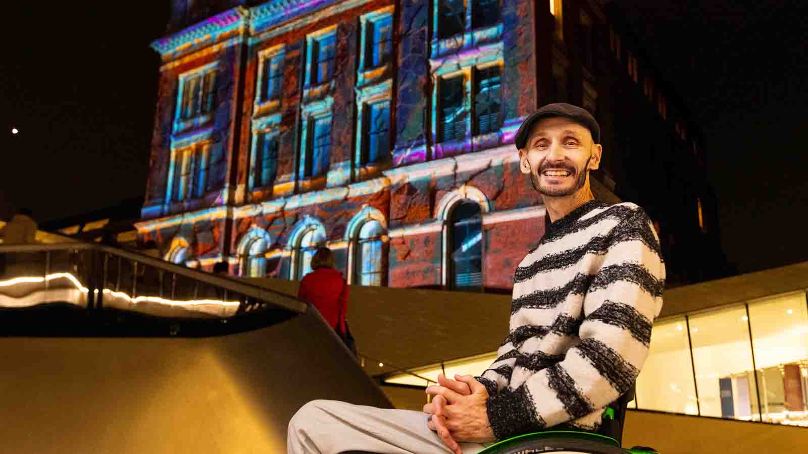 A man in a black flat cap and black and white striped jumper in a wheelchair (choreographer and producer Marc Brew) pictured in front of an old building with a colourful light show projected on to it.