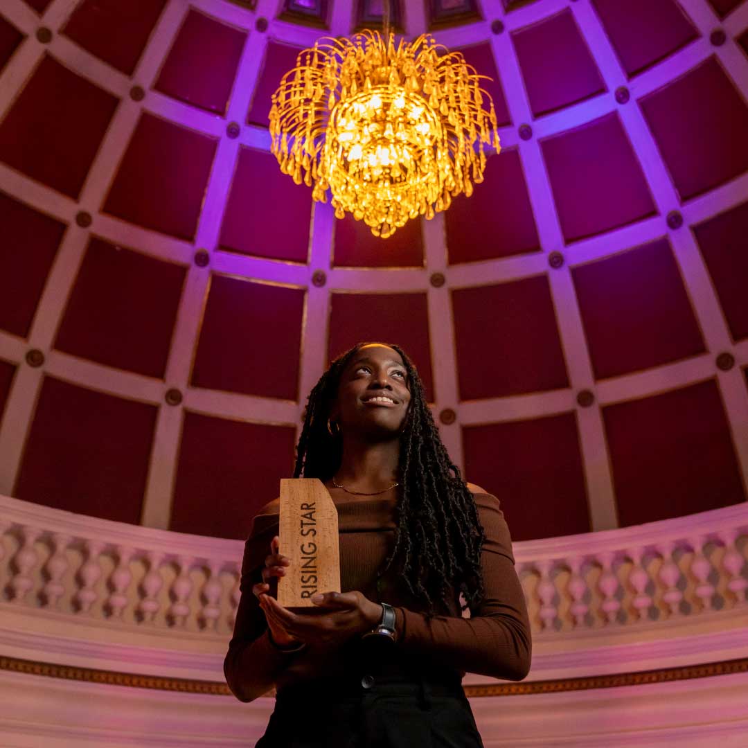 A Black woman with long dark hair styled in locks, wearing a gold necklace and soft red roll neck off shoulder sweater and holding a wooden award that reads ‘Rising Star’, this is Laura Oghagbon. She is standing in a large room with a chandelier above.