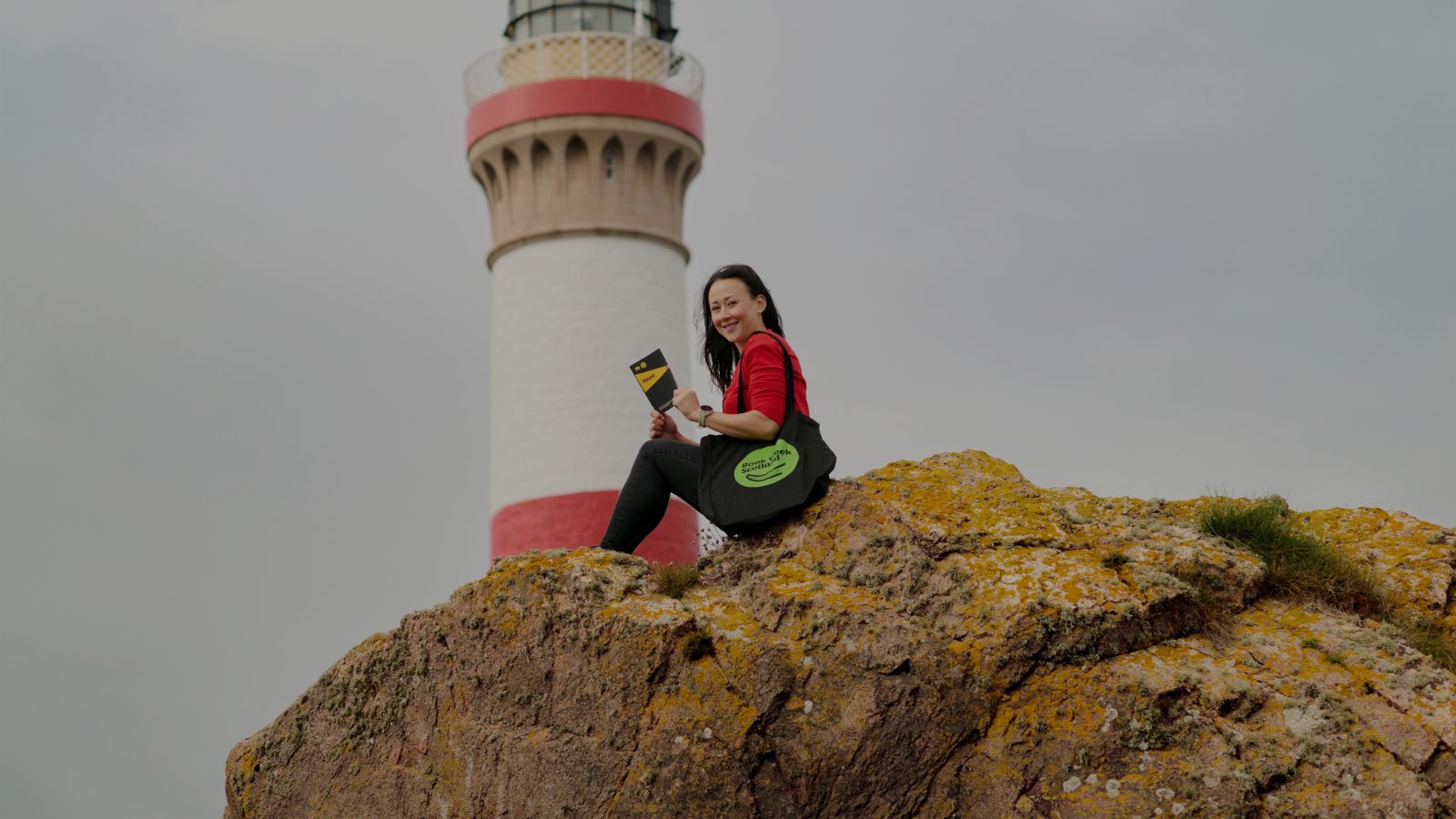 A woman sits on a large rock next to a lighthouse