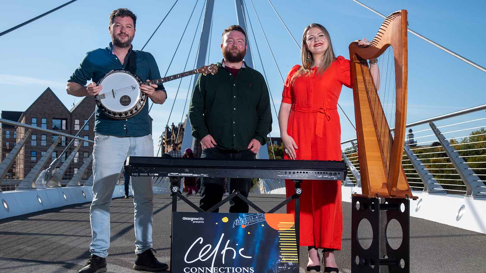 A man in a blue shirt holding a circular string instrument called a tenor banjo, this is Ciaran Ryan. Next to him in the middle is a man wearing a dark shirt and standing in front of a keyboard, this is Michael Biggins. On the end in a bright orange flow jumpsuit is a woman with long brown hair, standing with her left hand on a harp, this is Ciorstaidh Beaton. They are standing on a bridge outside on a beautiful bright sunny day. In front of them is a sign that reads 'Celtic Connections'