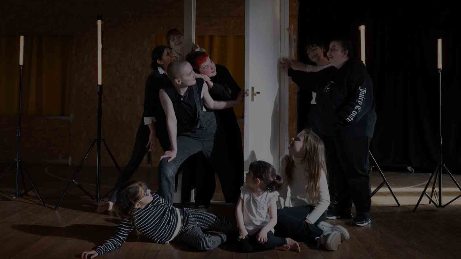 A group of young people performing together on a stage. Some younger members of the group are sitting on the floor, looking up. Two of them are peering round from bending a door that’s just been opened with another two looking over their shoulders. On the other side, two others smile as their friends come into view. They’re all dressed casually in comfy-looking, black and white clothes.