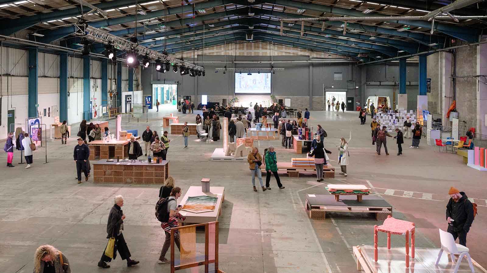 A large warehouse space with exposed walls, roof and cement floor. People milling about various displays with colourful and abstract pieces of furniture, clothing, sculpture and artwork. This is Dundee Design Festival.