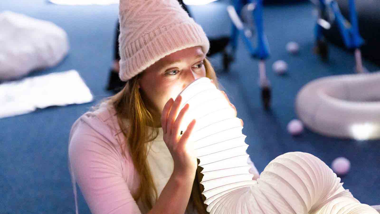 A woman wearing a pastel-pink winter bobble hat and pink top holds a white condenser vent up over her mouth and nose - the vent appears lit from inside and glows softly over her face and hands.