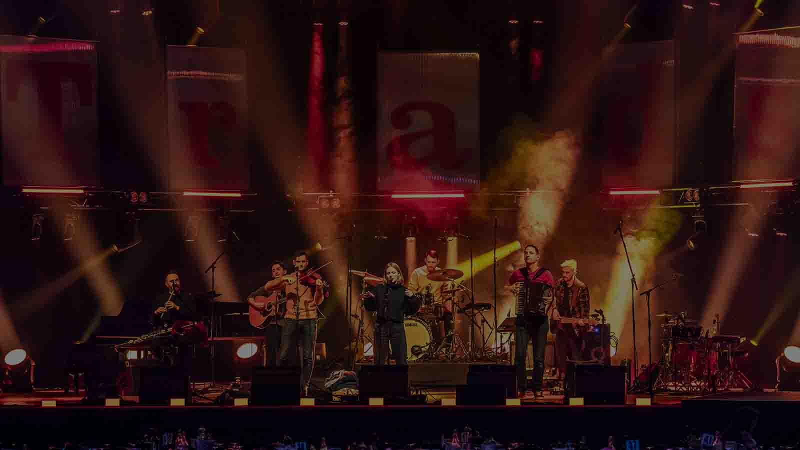 A stage lit up in bright, vibrant red and yellow lights. A group of musicians are performing together - a singer, an accordionist, a violinist, two guitarists, a flautist and a drummer. This is Mànran at the 2023 Trad music awards  ceremony.