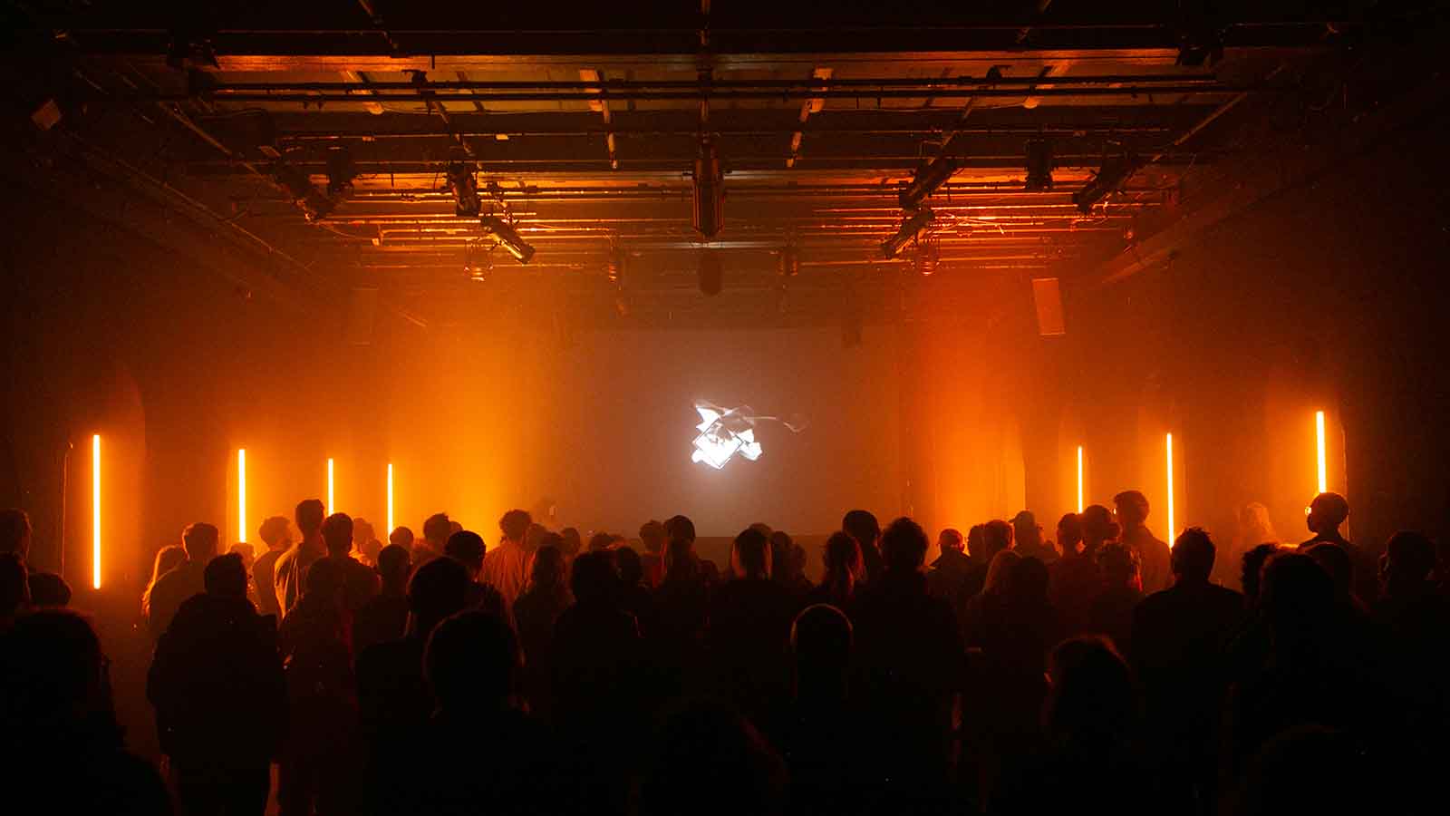 A crowd of people in a darkened room bathed in warm orange light emanating from several thin vertical lights on either side of the room. On the back wall a black and white image is displayed.