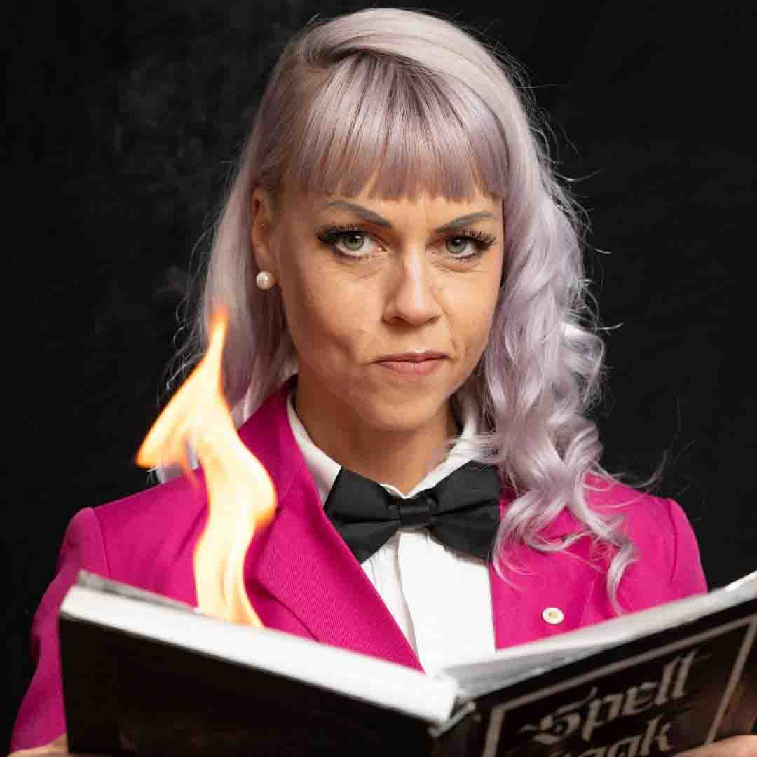 A woman with pale lilac/white hair wearing a formal white shirt and black bow tie and a cropped, shocking pink blazer. She is holding an open a ‘spell book’ which produces a bright yellow flame, as if by magic. This is Louise Andree