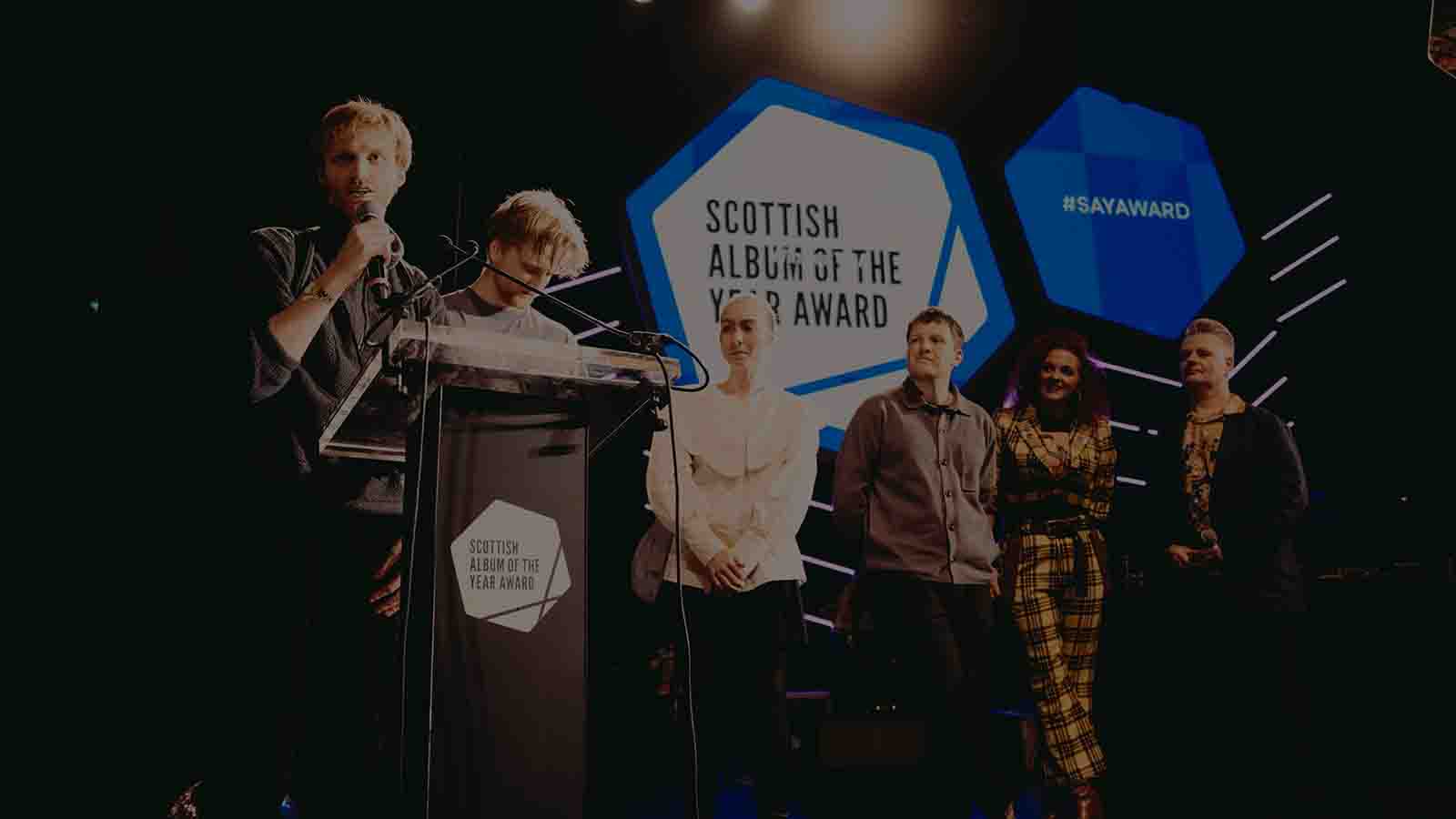 A man standing at a lectern on a stage speaks into a microphone. Next to him are more people, lit by stage lights, smiling and watching the speaker. These are the members of band rEDOLENT, accepting the Scottish Album of the Year award on stage.