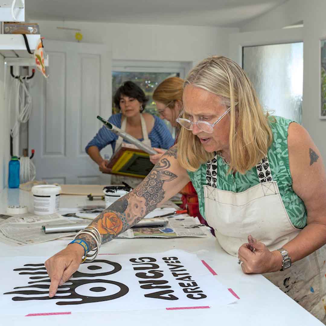 Three people wearing aprons stand at a long-bench doing screen-printing. The person in the foreground, with shoulder-length blonde hair, glasses and a sleeve of tattoos, works on a piece that features the Angus Creatives logo.